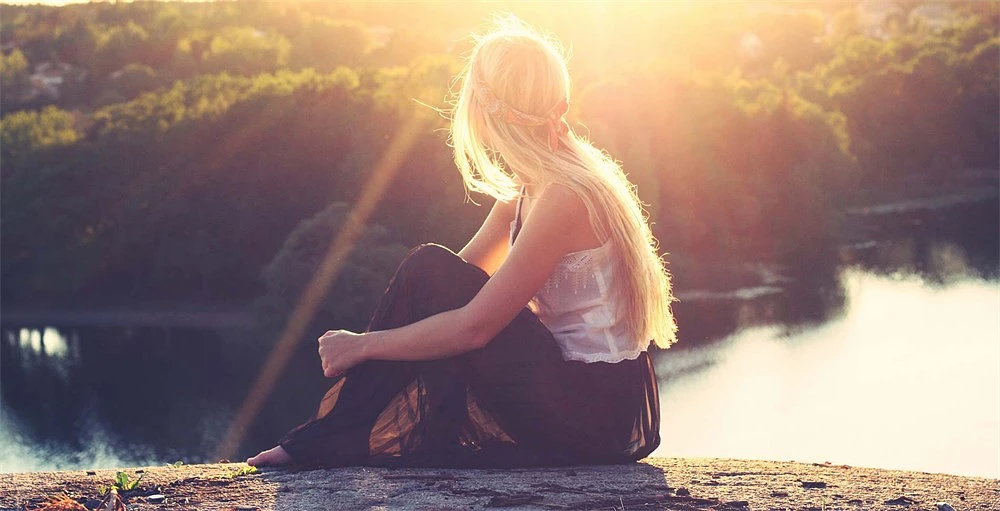 A student sits on a rock overlooking a lake and an area of trees with the sun shining on her