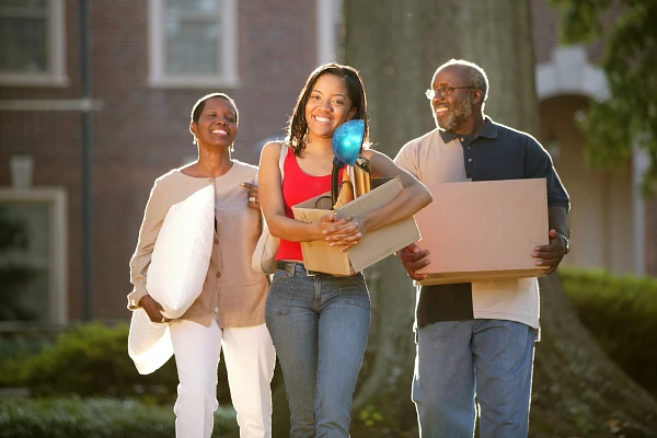 A young woman, smiling and carrying items, walks with two adults, possibly her parents, who are also carrying belongings.
