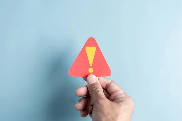 A hand is holding a red triangular warning sign with an exclamation mark on a blue background.