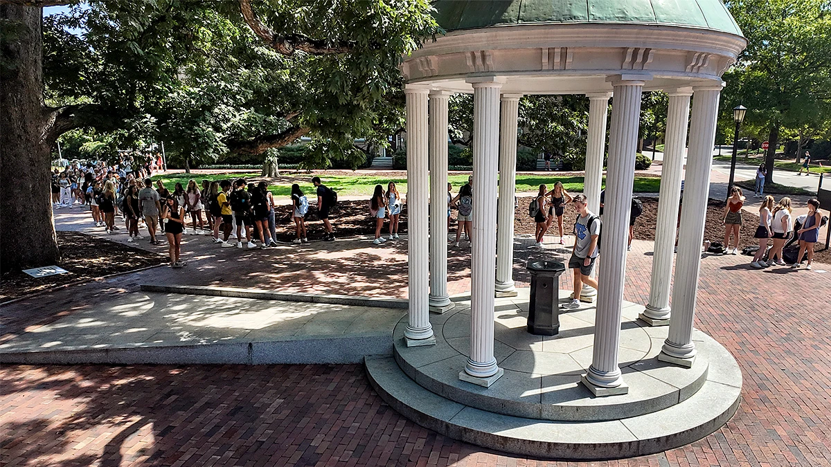 A group of people, mostly young adults, congregate near a white columned pavilion structure in a shaded outdoor area.