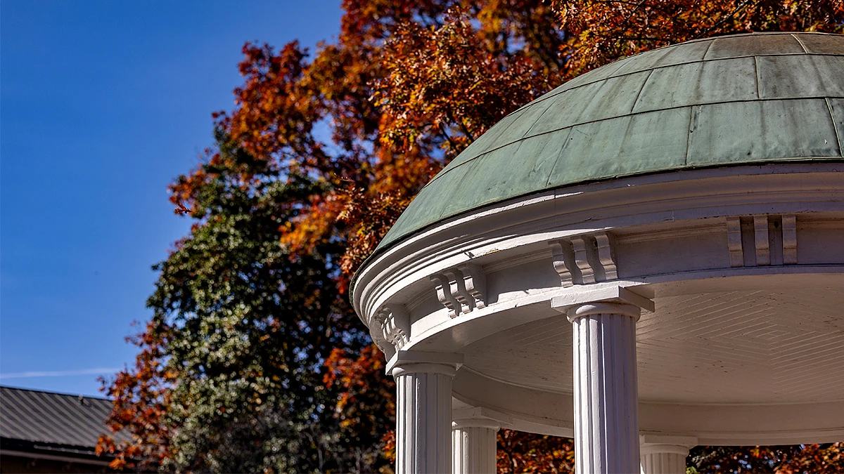 A white, columned gazebo with a green dome is set against a backdrop of trees with vibrant autumn foliage.
