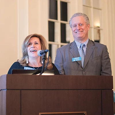 A woman and a man are standing at a podium, smiling, with a microphone in front of them at The Carolina Inn.