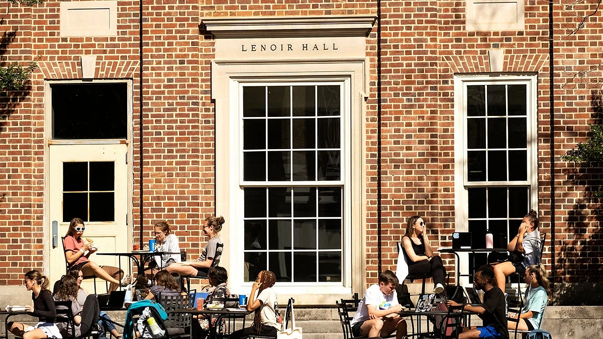 A group of people are sitting and studying or socializing outside Lenoir Hall on a sunny day.