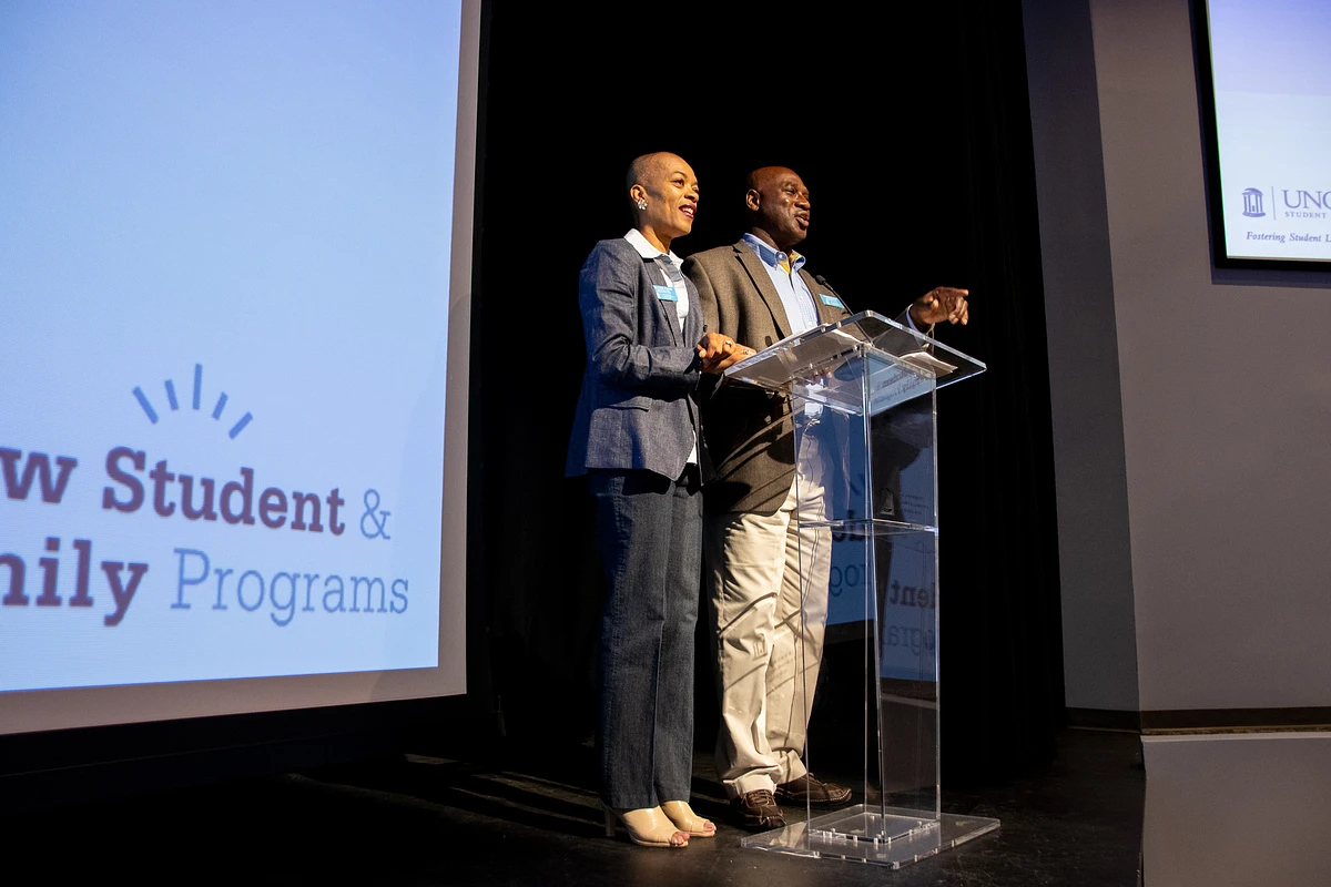 Two people are speaking at a podium during a presentation for "New Student & Family Programs."